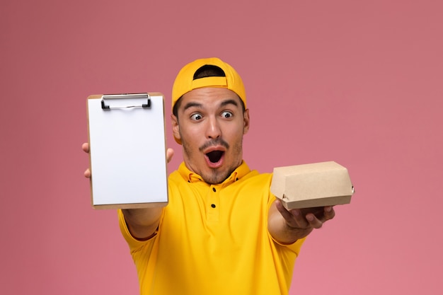 Mensajero masculino de vista frontal en uniforme amarillo con bloc de notas y pequeño paquete de comida sobre fondo rosa claro.