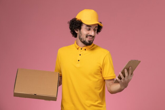 Mensajero masculino de vista frontal en uniforme amarillo con caja de entrega de alimentos y bloc de notas sonriendo en la pared rosa