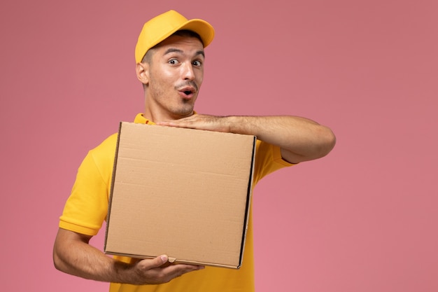 Mensajero masculino de vista frontal en uniforme amarillo con caja de entrega de alimentos con expresión divertida en el escritorio rosa