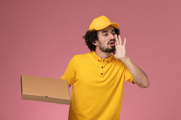 Mensajero masculino de vista frontal en uniforme amarillo con caja de entrega de alimentos llamando en la pared de color rosa claro