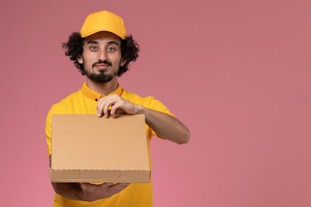 Mensajero masculino de vista frontal en uniforme amarillo con caja de entrega de alimentos en la pared de color rosa claro