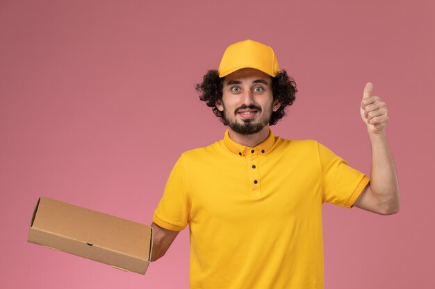 Mensajero masculino de vista frontal en uniforme amarillo con caja de entrega de alimentos en la pared de color rosa claro