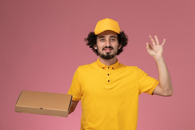 Mensajero masculino de vista frontal en uniforme amarillo con caja de entrega de alimentos en la pared de color rosa claro