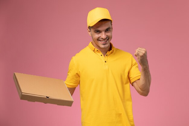 Mensajero masculino de vista frontal en uniforme amarillo con caja de entrega de alimentos y regocijo en el fondo rosa