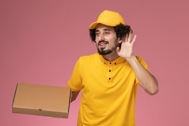 Mensajero masculino de vista frontal en uniforme amarillo con caja de entrega de alimentos tratando de escuchar en la pared de color rosa claro