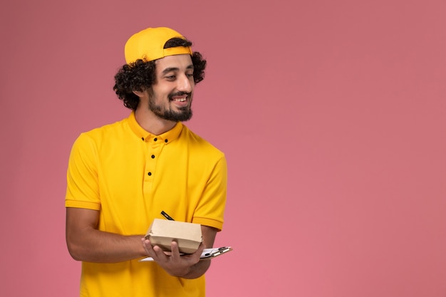 Mensajero masculino de vista frontal en uniforme amarillo y capa con bloc de notas y pequeño paquete de comida de entrega en sus manos escribiendo notas sobre fondo rosa.