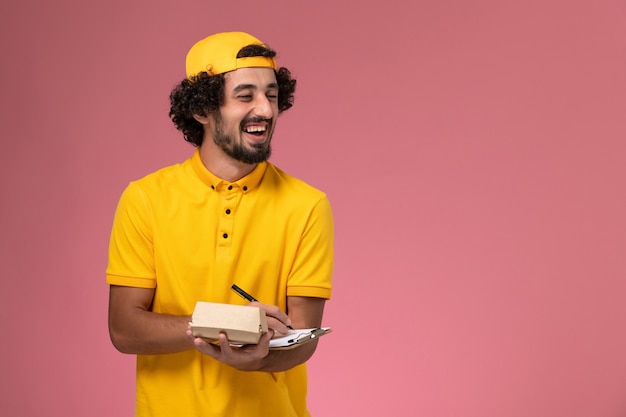 Mensajero masculino de vista frontal en uniforme amarillo y capa con bloc de notas y pequeño paquete de comida de entrega en sus manos escribiendo notas sobre fondo rosa.