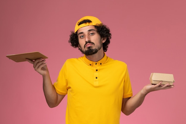 Mensajero masculino de vista frontal en uniforme amarillo y capa con bloc de notas y pequeño paquete de comida de entrega en sus manos sobre fondo rosa.