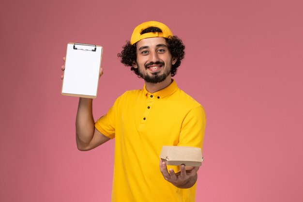 Mensajero masculino de vista frontal en uniforme amarillo y capa con bloc de notas y pequeño paquete de comida de entrega en sus manos sobre fondo rosa.