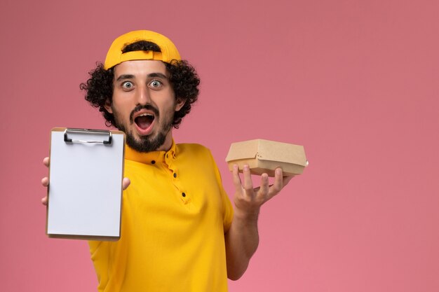 Mensajero masculino de vista frontal en uniforme amarillo y capa con bloc de notas y pequeño paquete de comida de entrega en sus manos sobre fondo rosa.