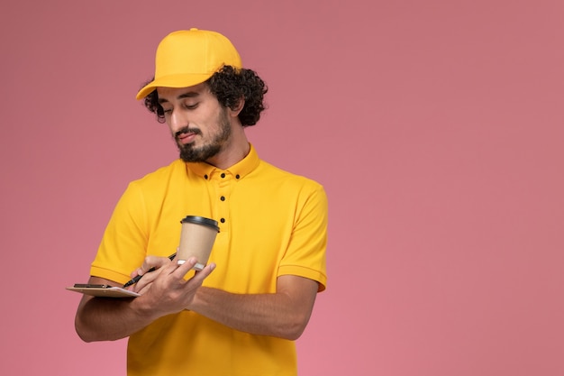 Foto gratuita mensajero masculino de vista frontal en uniforme amarillo y capa con bolígrafo de taza de café de entrega y bloc de notas en la pared rosa