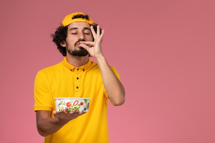 Foto gratuito mensajero masculino de vista frontal en uniforme amarillo y capa con cuenco de entrega redondo en sus manos posando sobre el fondo rosa.