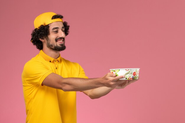 Mensajero masculino de vista frontal en uniforme amarillo y capa con cuenco de entrega redondo en sus manos sobre el fondo rosa claro.