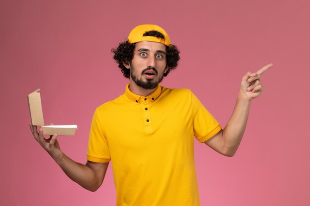 Mensajero masculino de vista frontal en uniforme amarillo y capa con paquete de comida de entrega pequeño abierto en sus manos sobre fondo rosa claro.