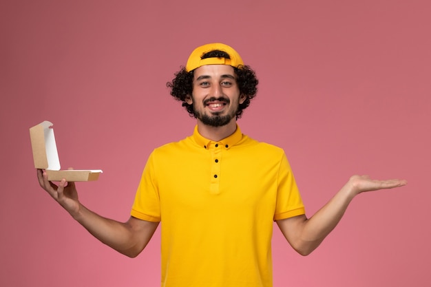 Mensajero masculino de vista frontal en uniforme amarillo y capa con paquete de comida de entrega pequeño abierto en sus manos sobre fondo rosa.