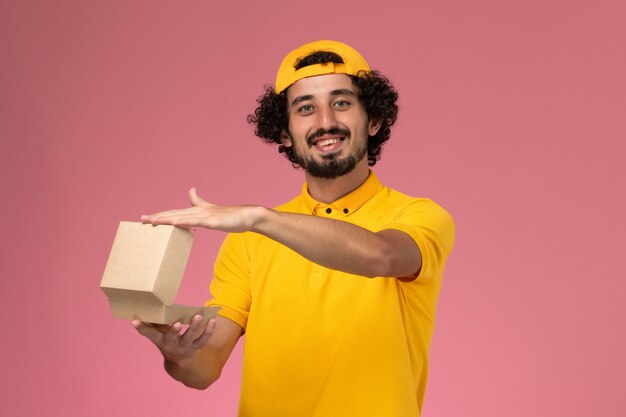 Mensajero masculino de vista frontal en uniforme amarillo y capa con paquete de comida de entrega pequeño abierto en sus manos sobre fondo rosa.