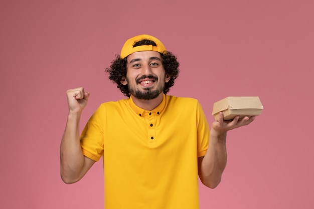 Mensajero masculino de vista frontal en uniforme amarillo y capa con pequeño paquete de comida de entrega en sus manos regocijándose en el fondo rosa.