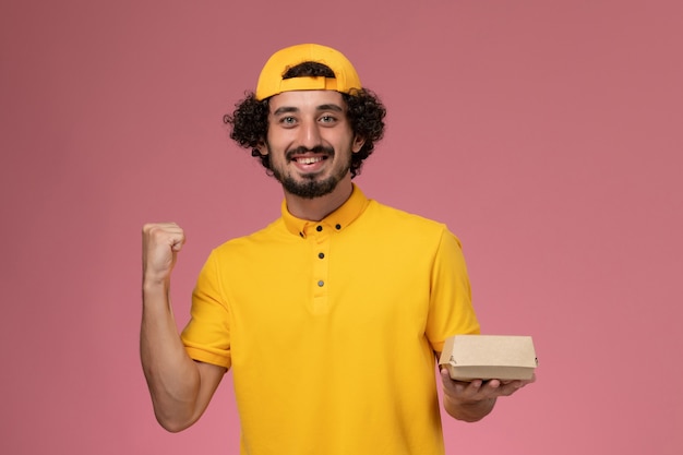 Mensajero masculino de vista frontal en uniforme amarillo y capa con pequeño paquete de comida de entrega en sus manos regocijándose en el fondo rosa.
