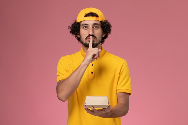 Mensajero masculino de vista frontal en uniforme amarillo y capa con pequeño paquete de comida de entrega en sus manos sobre el fondo rosa.