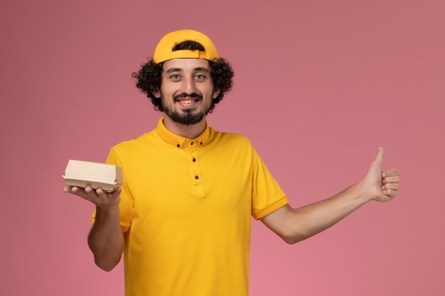 Mensajero masculino de vista frontal en uniforme amarillo y capa con pequeño paquete de comida de entrega en sus manos sobre el fondo rosa.