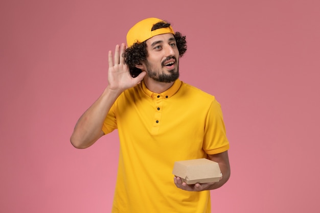 Mensajero masculino de vista frontal en uniforme amarillo y capa con pequeño paquete de comida de entrega en sus manos sobre el fondo rosa.