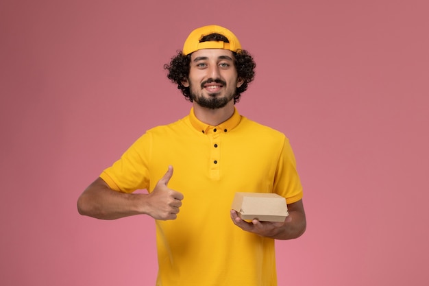 Mensajero masculino de vista frontal en uniforme amarillo y capa con pequeño paquete de comida de entrega en sus manos sobre el fondo rosa.