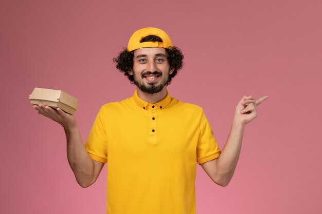 Mensajero masculino de vista frontal en uniforme amarillo y capa con pequeño paquete de comida de entrega en sus manos sobre el fondo rosa.