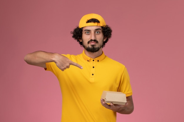 Mensajero masculino de vista frontal en uniforme amarillo y capa con pequeño paquete de comida de entrega en sus manos sobre el fondo rosa.