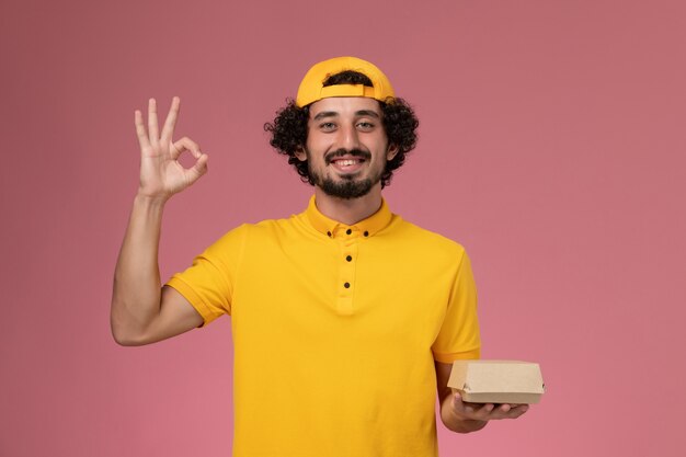 Mensajero masculino vista frontal en uniforme amarillo y capa con pequeño paquete de comida de entrega en sus manos sonriendo sobre el fondo rosa