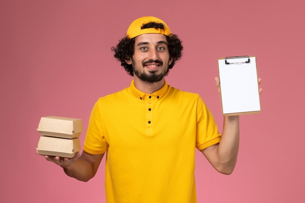 Mensajero masculino de vista frontal en uniforme amarillo y capa con pequeños paquetes de comida de entrega y bloc de notas en sus manos sobre el fondo rosa claro.