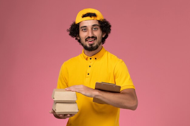 Mensajero masculino de vista frontal en uniforme amarillo y capa con pequeños paquetes de comida de entrega y bloc de notas en sus manos sobre el fondo rosa claro.