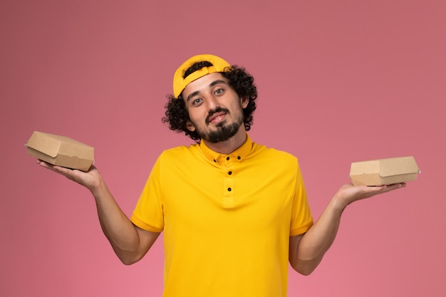 Mensajero masculino de vista frontal en uniforme amarillo y capa con pequeños paquetes de comida de entrega en sus manos sobre el fondo rosa.