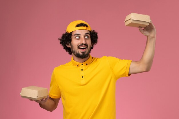 Mensajero masculino de vista frontal en uniforme amarillo y capa con pequeños paquetes de comida de entrega en sus manos sobre el fondo rosa.