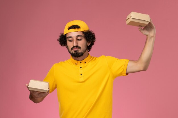 Mensajero masculino de vista frontal en uniforme amarillo y capa con pequeños paquetes de comida de entrega en sus manos sobre el fondo rosa.