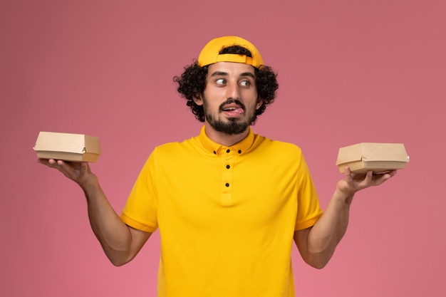 Mensajero masculino de vista frontal en uniforme amarillo y capa con pequeños paquetes de comida de entrega en sus manos sobre el fondo rosa.