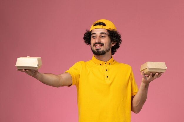 Mensajero masculino de vista frontal en uniforme amarillo y capa con pequeños paquetes de comida de entrega en sus manos sonriendo sobre el fondo rosa.