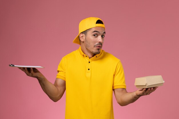 Mensajero masculino de la vista frontal en uniforme amarillo y capa que sostiene el pequeño bloc de notas de paquete de comida de entrega sobre fondo rosa