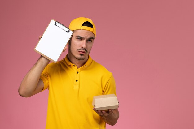 Mensajero masculino de la vista frontal en uniforme amarillo y capa que sostiene el pequeño paquete de comida de entrega y el bloc de notas con expresión de pensamiento en el escritorio rosa.