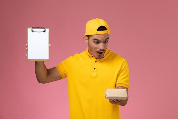 Mensajero masculino de la vista frontal en uniforme amarillo y capa que sostiene el pequeño paquete de comida de entrega y el bloc de notas con expresión de sorpresa sobre fondo rosa.