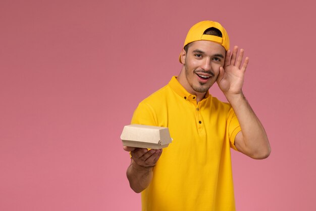 Mensajero masculino de la vista frontal en uniforme amarillo y capa que sostiene el pequeño paquete de comida de entrega en el fondo rosa claro.