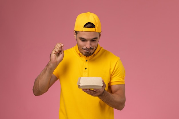 Foto gratuita mensajero masculino de la vista frontal en uniforme amarillo y capa que sostiene el pequeño paquete de comida de entrega en el fondo rosa claro.