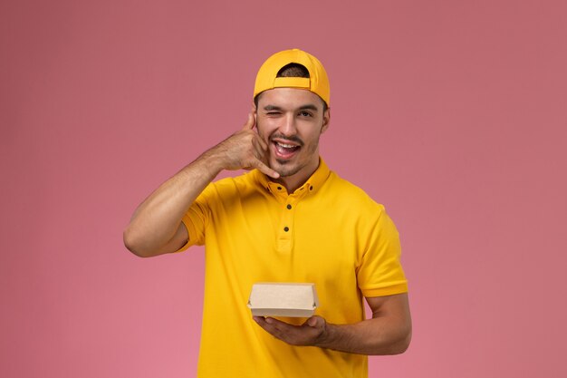 Mensajero masculino de la vista frontal en uniforme amarillo y capa que sostiene el pequeño paquete de comida de entrega en el fondo rosa.