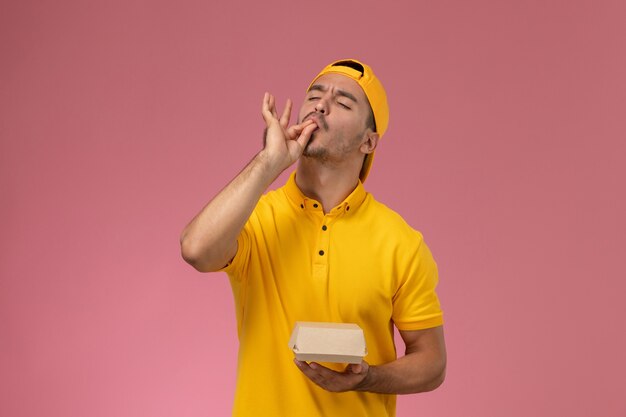 Mensajero masculino de la vista frontal en uniforme amarillo y capa que sostiene el pequeño paquete de comida de entrega en el fondo rosa.