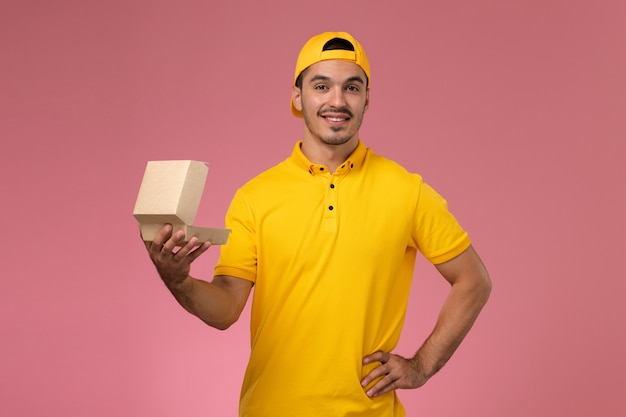 Mensajero masculino de vista frontal en uniforme amarillo y capa sosteniendo y abriendo pequeño paquete de comida de entrega sonriendo sobre el fondo rosa.