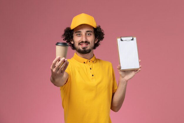 Mensajero masculino de vista frontal en uniforme amarillo y capa sosteniendo la taza de café de entrega y el bloc de notas en la pared rosa