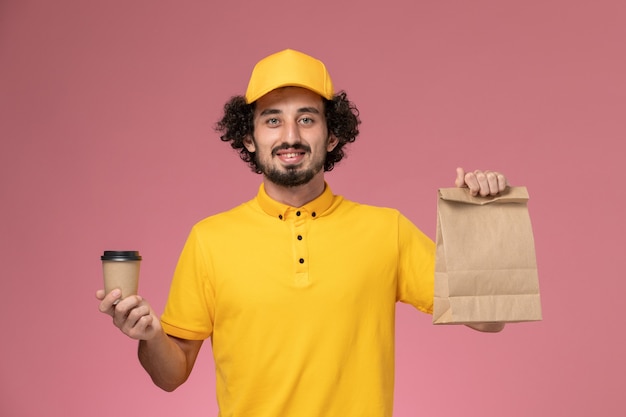 Mensajero masculino de vista frontal en uniforme amarillo y capa sosteniendo la taza de café de entrega y el paquete de comida en el escritorio rosa uniforme trabajo empresa de servicio trabajador masculino