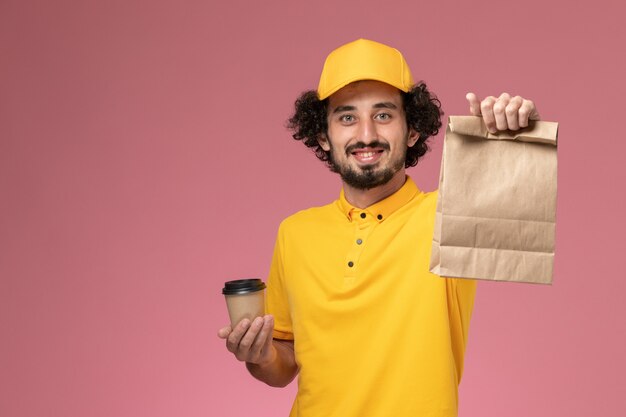 Mensajero masculino de vista frontal en uniforme amarillo y capa sosteniendo la taza de café de entrega y el paquete de comida en el escritorio rosa uniforme trabajo empresa trabajador masculino