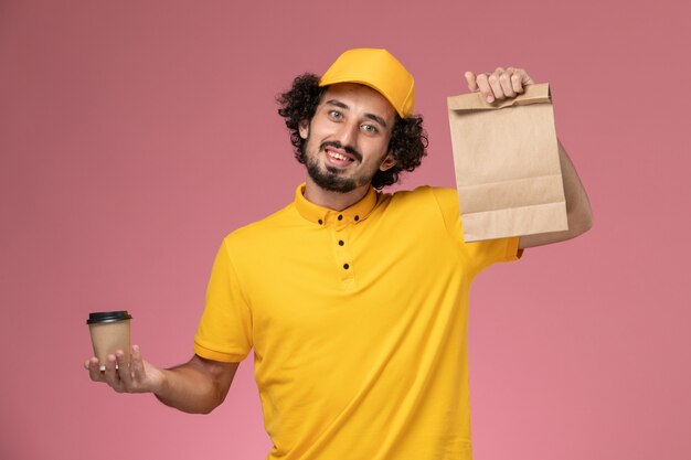 Mensajero masculino de vista frontal en uniforme amarillo y capa sosteniendo la taza de café de entrega y el paquete de comida en el uniforme de escritorio rosa empresa de servicio masculino