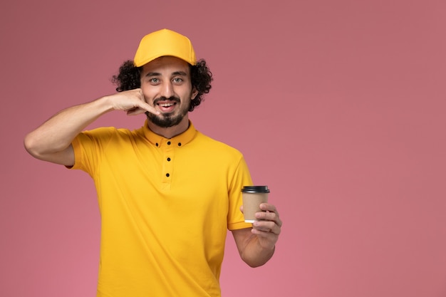 Mensajero masculino de vista frontal en uniforme amarillo y capa sosteniendo la taza de café de entrega en la pared de color rosa claro