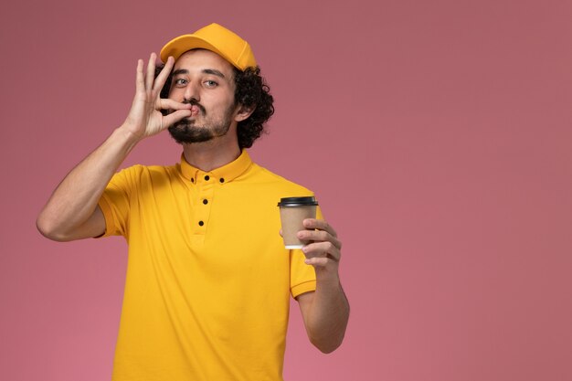 Mensajero masculino de vista frontal en uniforme amarillo y capa sosteniendo la taza de café de entrega en la pared rosa